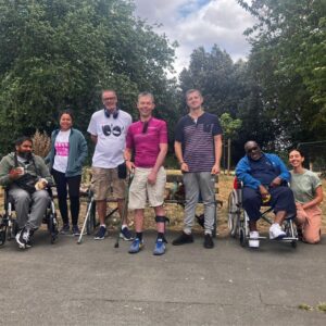 A group of members, staff and volunteers from Headway East London are in a park on a summer day. Two of the members use wheelchairs, two use walking sticks. They are all smiling,