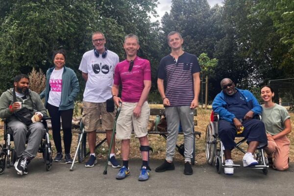 A group of members, staff and volunteers from Headway East London are in a park on a summer day. Two of the members use wheelchairs, two use walking sticks. They are all smiling,