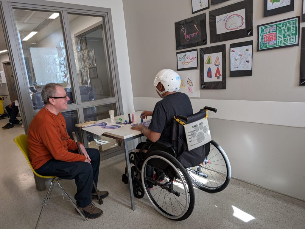 Two people in a room with a table and artwork on the wall. One person is a man in his sixties, smiling and looking at the table. The other person is visible only from the back, They are sitting in a wheelchair and wearing a protective white helmet. They are using a blue coloured pen to complete a drawing.