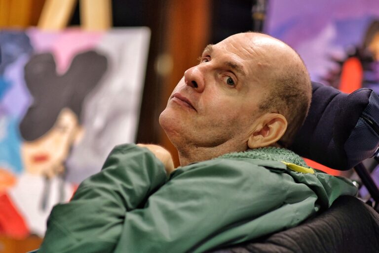 In the foreground is Jon, a member of Headway East London, who is sat in his wheelchair. He is a middle aged white man with a bald head, and he is wearing a green jumper. He is looking over his shoulder with a curious, soft expression and his head tilted to one side. In the background is one of his paintings in greys, pinks and blacks.
