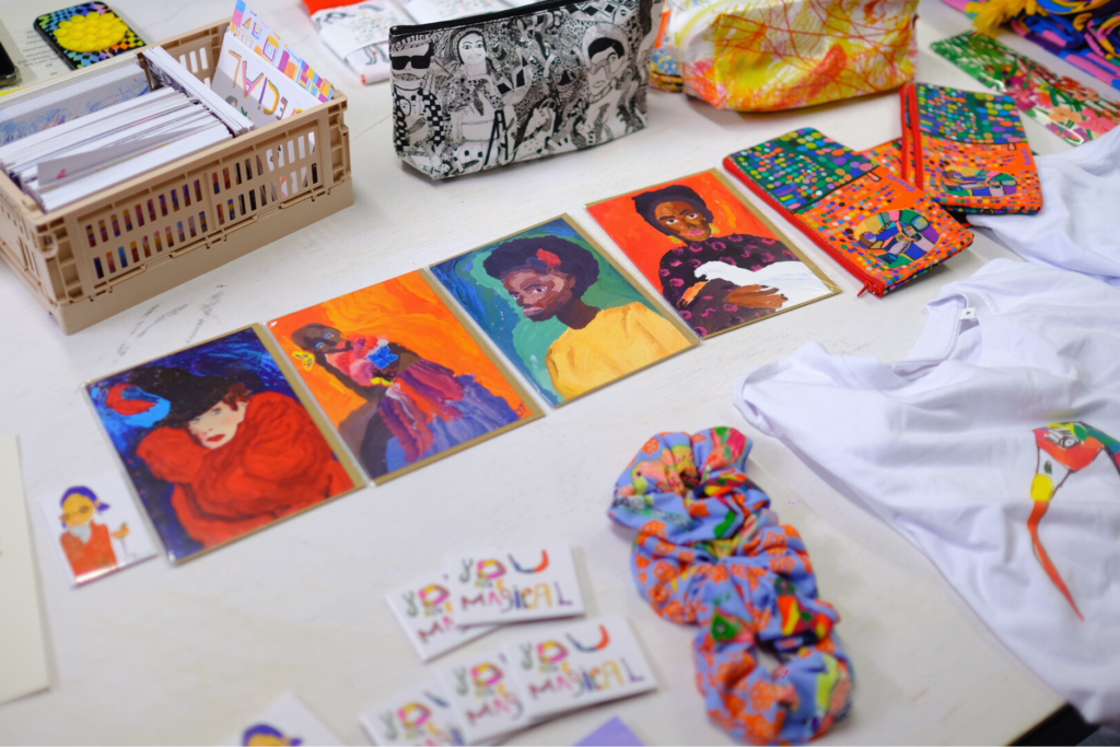 A picture of our merchandise table at one of our events. There are greeting cards, scrunchies, t-shorts, make-up bags and magnets. Everything is very colourful and laid out nicely.