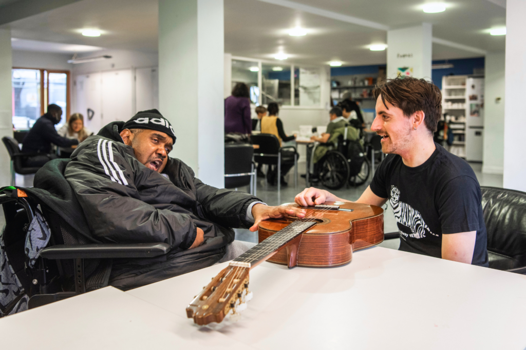 Two people stit in front of each other. In the middle there is a guitar on a table. They are both touching the guitar. The man on the right is a member of Headway East London. He is a Black man in his sixties. He is sitting on a wheelchair and singing. The man on the right is a staff member of Headway. He is a white man in his thirties. He is touching the chords of the guitar and smiling. In the background there is a rooom with tables and white columns.