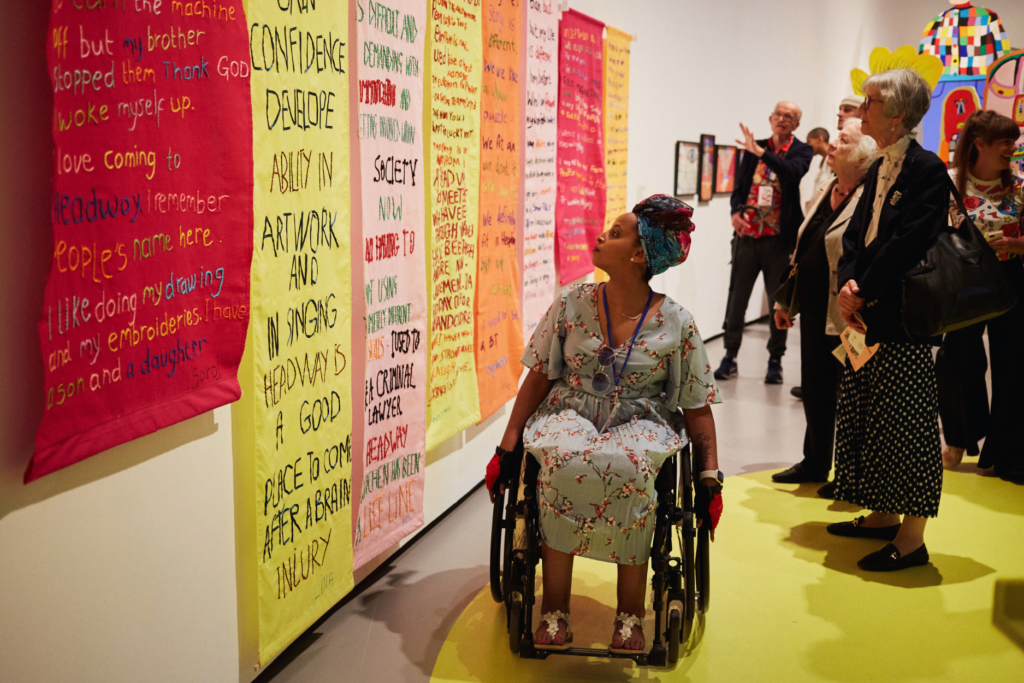 Our member Toki is in the Curve Galerry of the Barbican. She is an Ehtiopian woman in hher thirties. She is sitting on a wheelchair and she is wearing a dress with flower patterns. She is looking toward her right at a series of eight embroidered banners exhibited at the Barbican for our differently various exhibition. Each banner is different in colours and text. Each of them tells the story of a brain injury survivor.