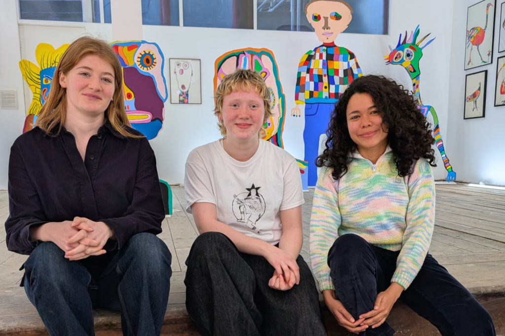 Three Headway East London volunteers sitting on the stairs at one our exhibitions. They are three girls looking at the camera and smiling. Behind them there are colourful giant figures from our artist Jason.