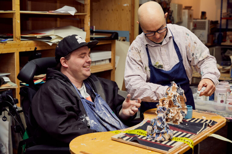 Two men in working together in the Headway East London art studio. One is a volunteer and one is a member of Headway. The volunteer is helping the member painting a ceramic sculpture that they have made togehter. The member is a white, young guy. He is sitting on a wheelchair, he is smiling and looking at the table in front of him. He is wearing an apron and a cap. The volunteer is a white man. He is standing, wearing an apron, black glasses and a white T-shirt. He is looking at the artwork. Make a donation so we can continue support people living with brain injury.