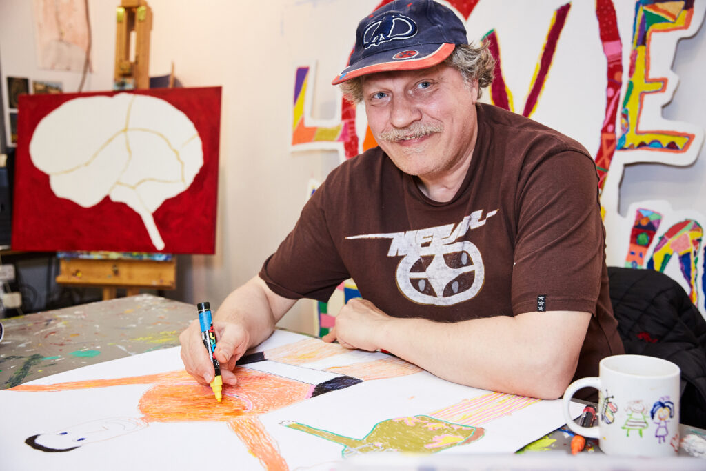 Tony Allen, a white man in his 50s with a blue cap, brown t-shirt and a friendly smile sits at a table with a yellow posca pen in his hand, working on a drawing. He is sat in the Submit to Love art studio, and behind him are colourful artworks arranged along the walls.