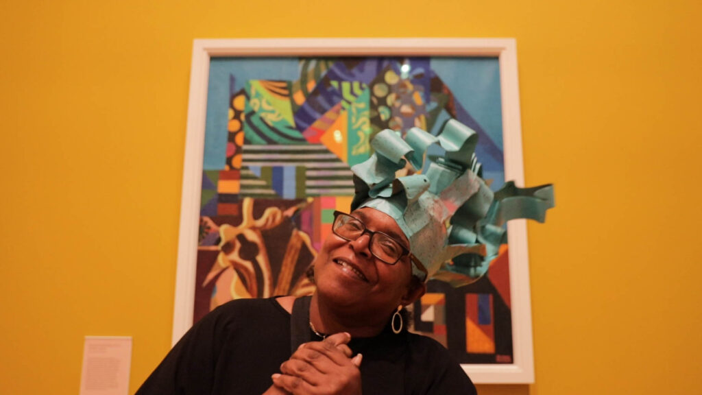Theresa, a member of Headway East London, is sitting in front of a colourful painting on a bright yellow wall at Whitechapel Gallery. She is a middle aged black woman with glasses, she is smiling and clasping her hands together. On her head is a fantastical hat made of blue curved paper, like a post-modern top hat, which she has hand-made.